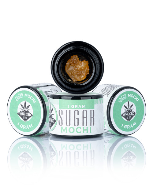Mochi Sugar (85.400% THCa) in a black container with visible golden crumble and green labeling. Mochi Sugar (85.400% THCa) in a black container with visible golden crumble and green labeling.