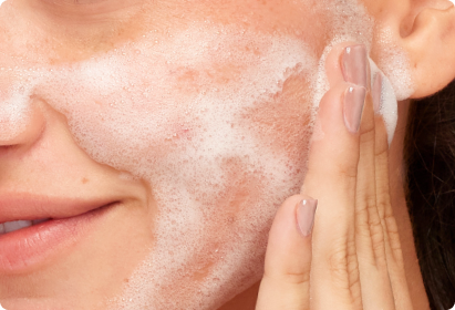 Close up image of a woman lathering cleansing foam on her left cheek.