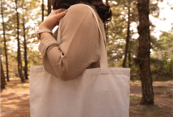 Person carrying a neutral canvas tote bag that has the Olay logo with a printed graphic that reads “Ageless Beauty” in a wooded outdoor setting. A red price badge reads $45 with $120+ value