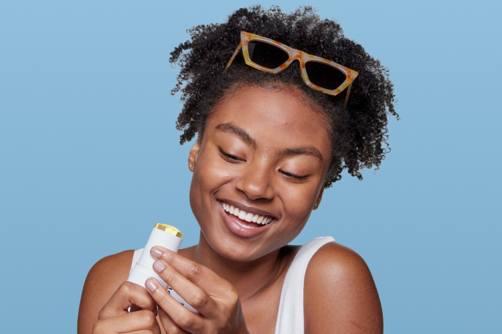 Smiling woman applying Olay Glow + Protect SPF 50 sunscreen stick with sunglasses on head, against a blue background