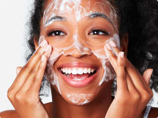 Young woman smiling while applying foamy cleanser to her face.