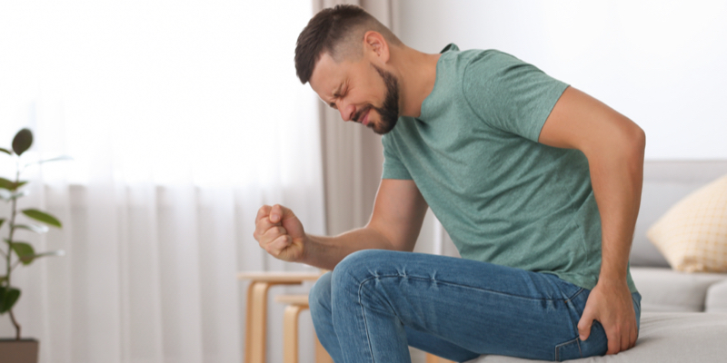 A man in a green shirt and jeans sits on a couchin discomfort from hemorrhoids. A plant, stool, and pillow are visible in the background of the home setting.