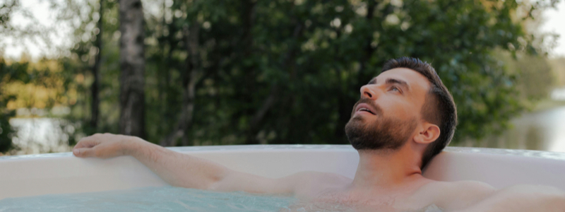 A person relaxes in an outdoor hot tub surrounded by trees and overlooking a body of water. The peaceful setting highlights comfort and wellness; the person's face is blurred for privacy.
