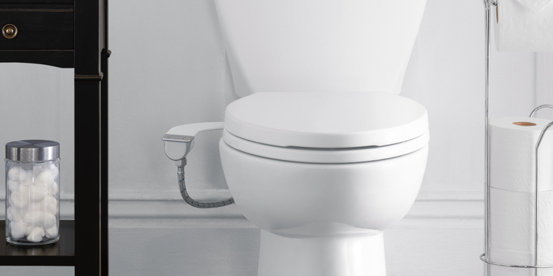 Modern white toilet with bidet attachment in a clean bathroom, flanked by a black shelf with cotton balls and a chrome stand holding toilet paper rolls.