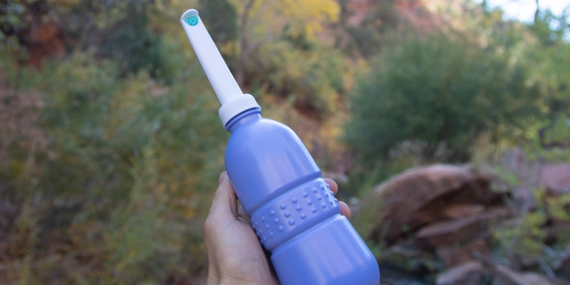 Hand holding a portable travel bidet with white nozzle and blue body, set against an outdoor background with greenery and rocks, highlighting its convenience for travel or nature use.