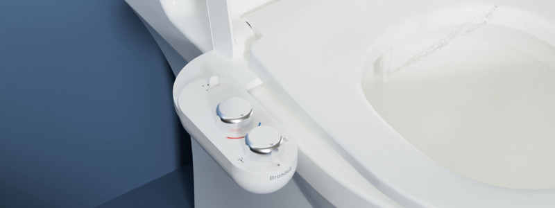 SimpleSpa Eco Advanced Warm Water Bidet Attachment installed on a toilet, showing dual control knobs and a warm-water spray stream inside the bowl.