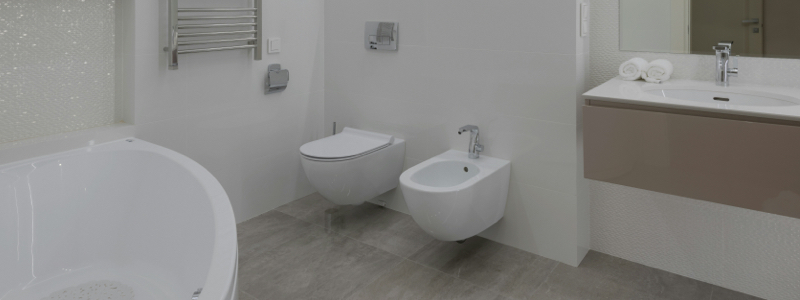 Modern bathroom with white tub, wall-mounted toilet and bidet, gray tile floor, towel rack, and sink with mirror and rolled white towels.