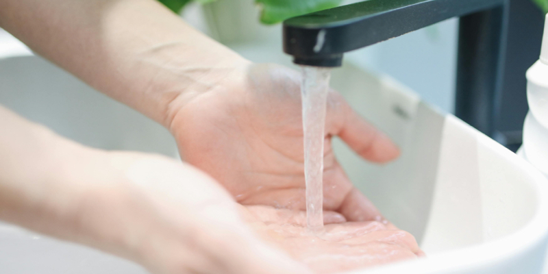 Close-up of hands being washed under running water from a black faucet in a white sink, with green plants visible in the background.
