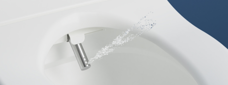 Close-up of a bidet nozzle extending from beneath a toilet seat, spraying a stream of water inside a clean white bowl.
