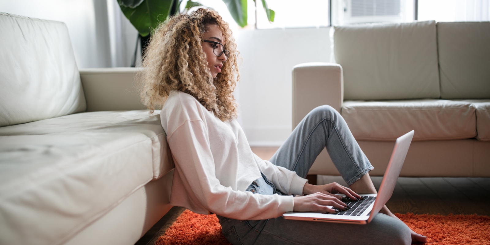 Woman student sitting on floor with laptop, leaning against bed.