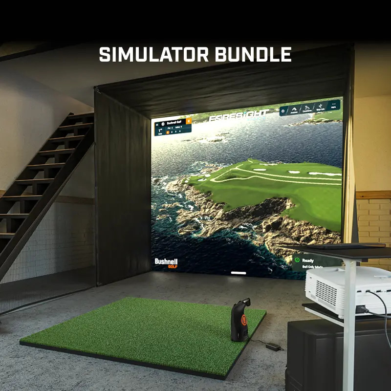 Indoor golf simulator setup featuring the LPI Simulator Bundle from Bushnell Golf, including a launch monitor, hitting mat, projector, and large impact screen displaying a realistic coastal golf course.