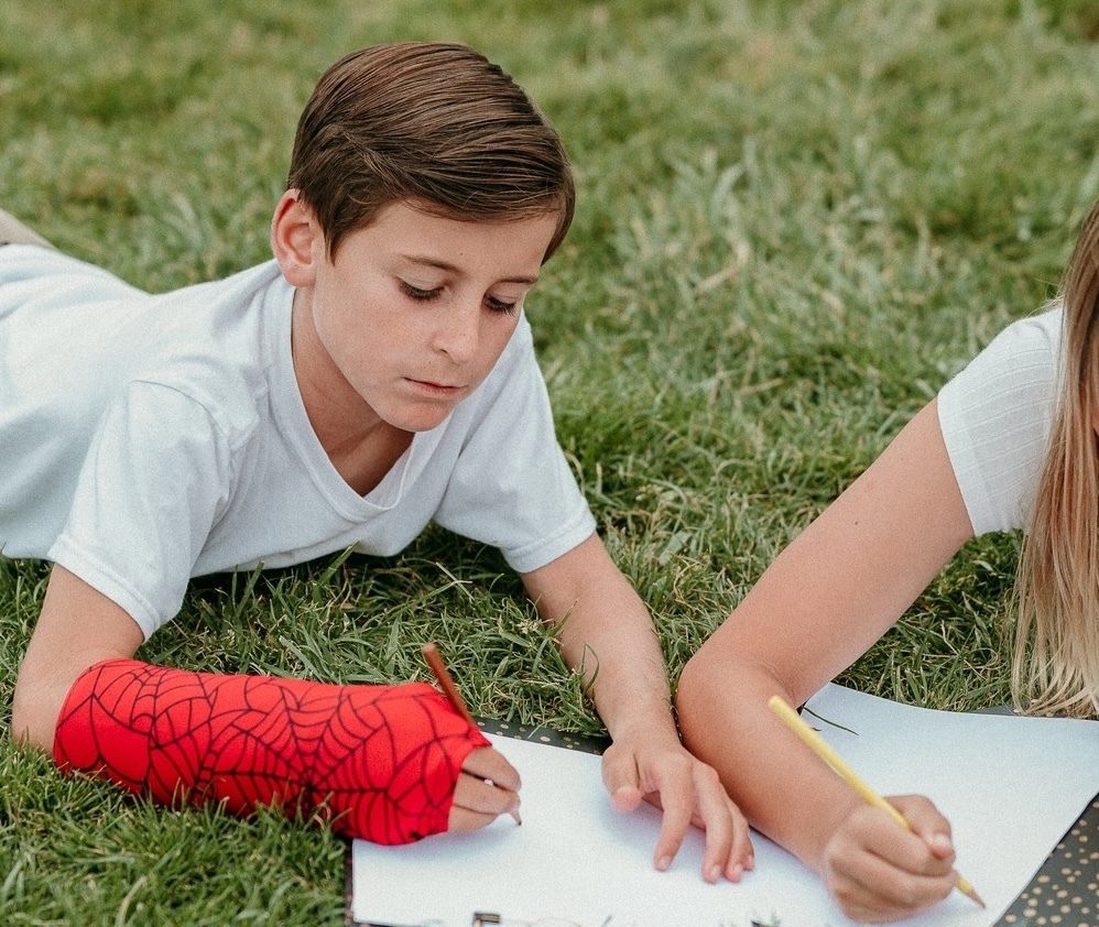 a boy wearing a red spider web arm cast coloring in the grass