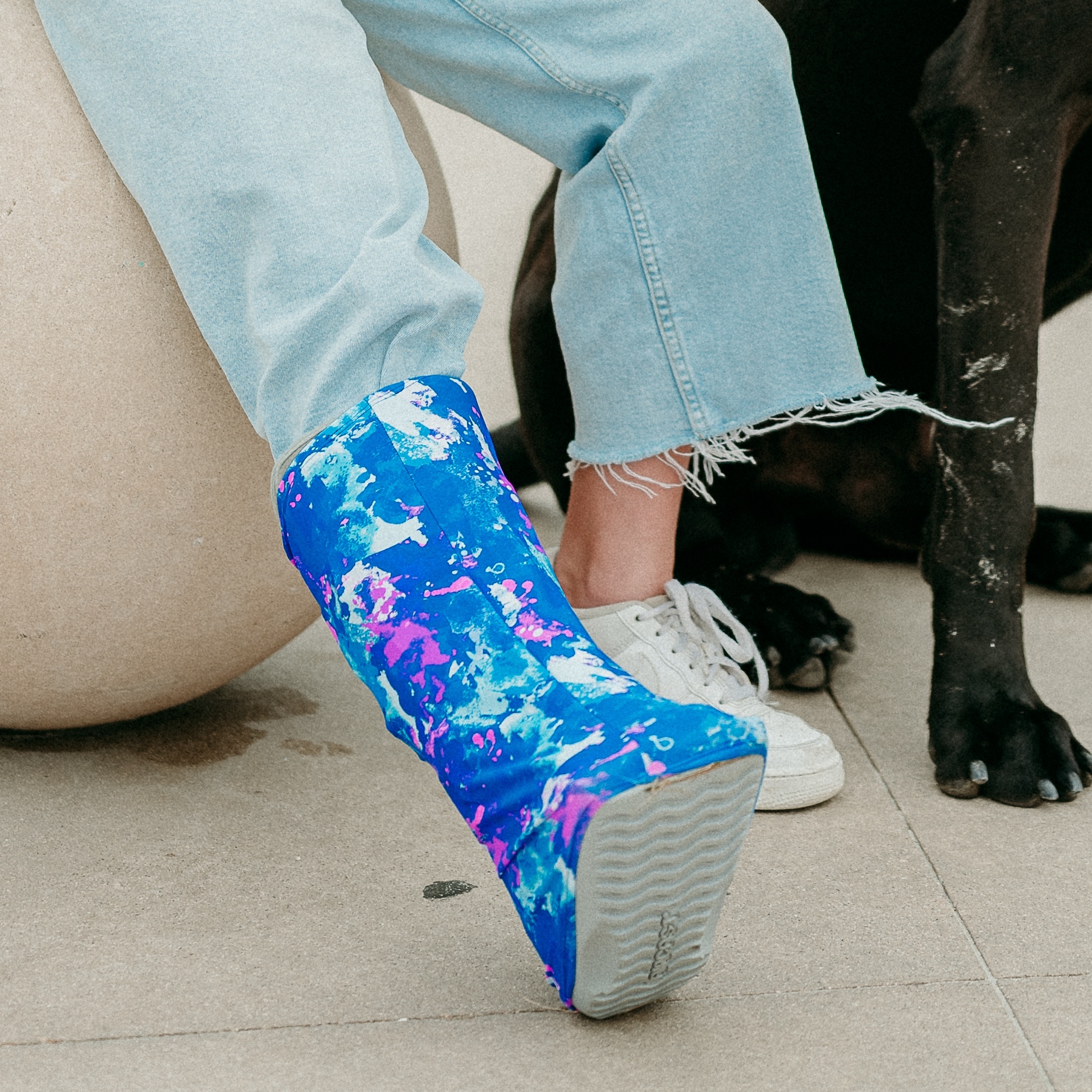 a girl wearing jeans, seen from the knee down wearing white sneakers and a colorful orthotic walking boot cover, black dog in background