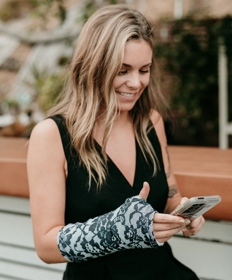 Smiling woman using her phone while wearing a black-and-white lace arm cast cover during recovery.