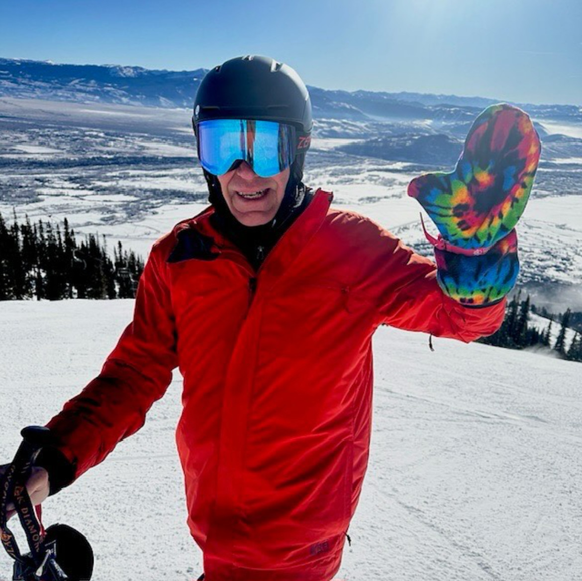 Person skiing on a snowy mountain wearing a colorful tie-dye recovery mitten while dressed in red ski gear and winter helmet.