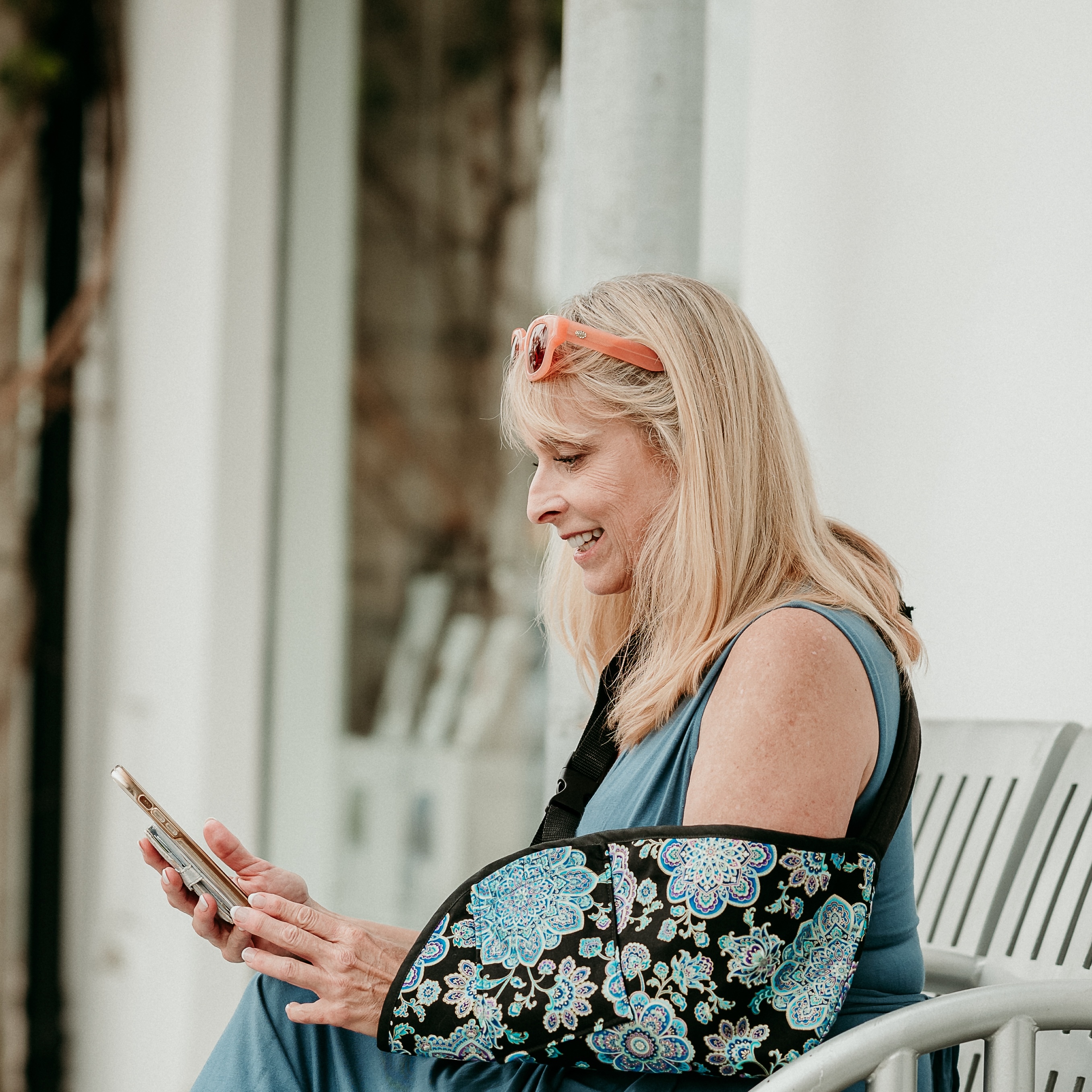 a woman with orange glasses on her head on her phone sitting on a bench wearing a paisley arm sling 