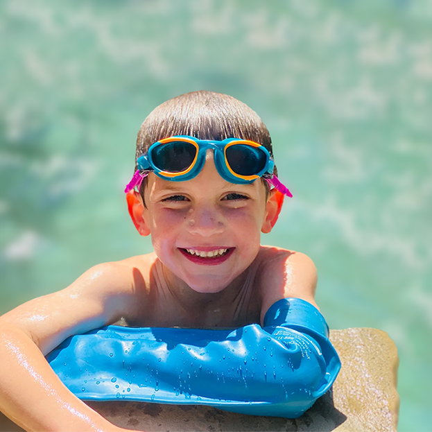 Smiling child wearing swim goggles and a blue waterproof arm cast cover while enjoying the water on a sunny day.
