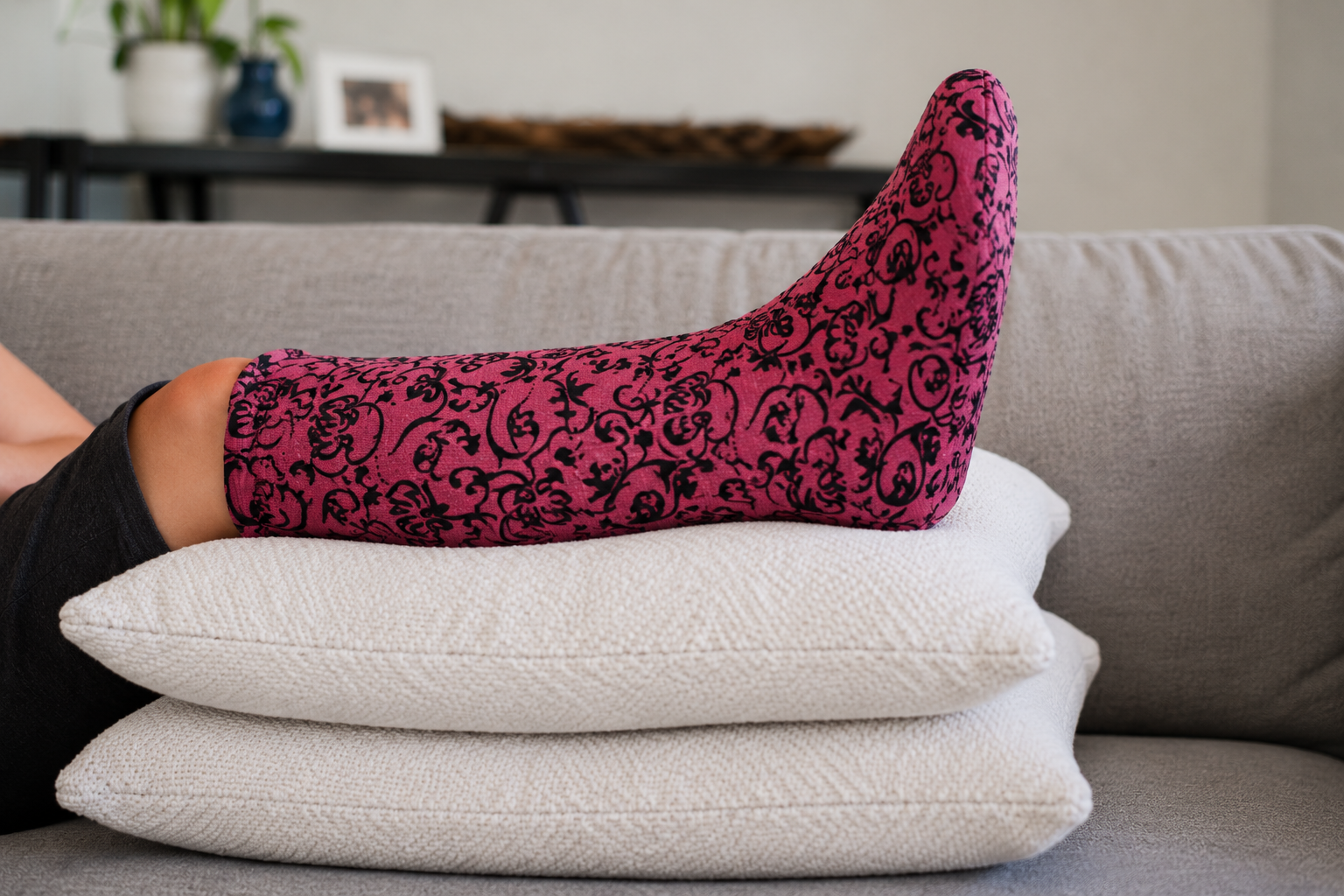 Person resting at home with a pink-and-black patterned leg cast cover elevated on pillows