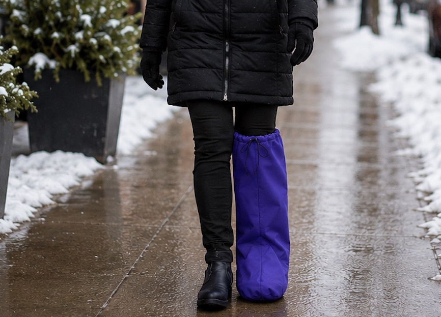Woman wearing a BootGuardzXtreme weather protection cover over an orthopedic walking boot while walking on a snowy city sidewalk. Weatherproof protection helps keep walking boots dry and protected from rain, snow, slush, and cold winter conditions.