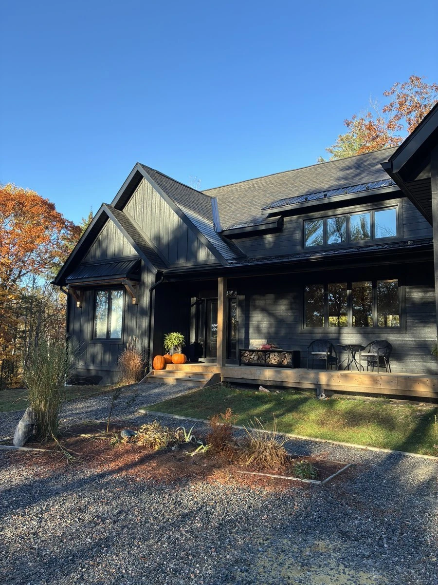 A dark-sided house with a covered front porch, stacked firewood and pumpkins by the steps, a gravel drive, and autumn trees under a clear blue sky