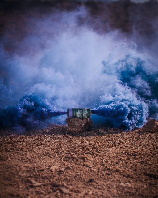 Blue smoke grenade lying on rocky dirt ground, blue smoke billowing outward on both sides in an open field