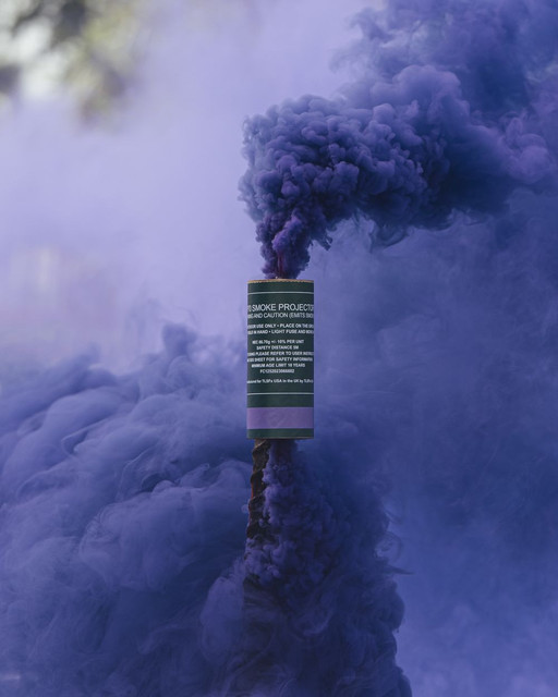 Purple smoke grenade planted upright, a dense purple column billowing upward against a blurred outdoor backdrop