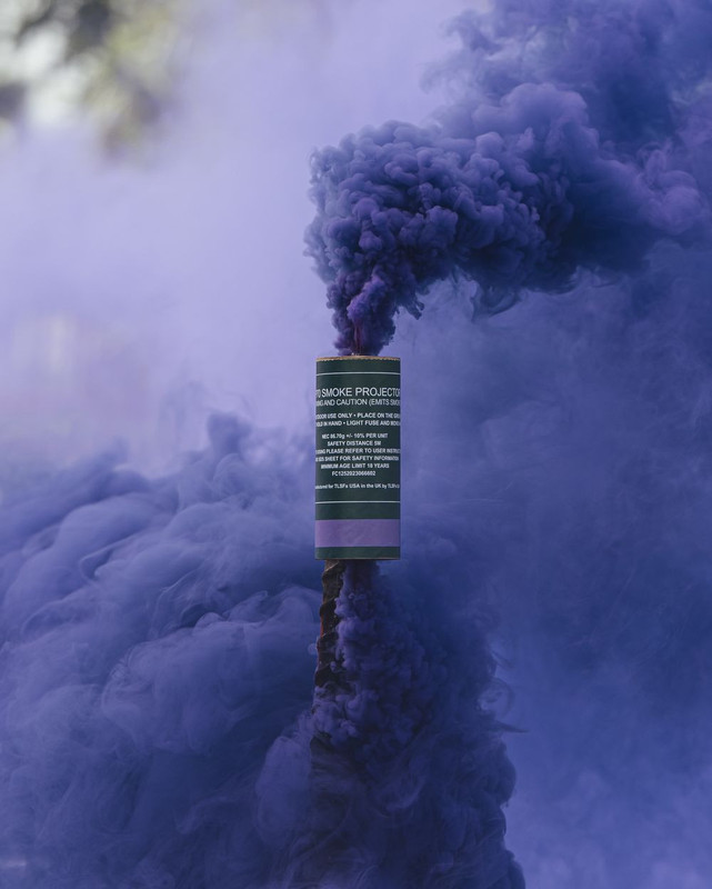 Purple smoke grenade planted upright, a dense purple column billowing upward against a blurred outdoor backdrop