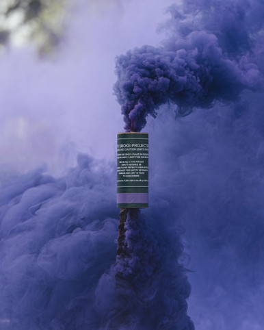 Purple smoke grenade planted upright, a dense purple column billowing upward against a blurred outdoor backdrop