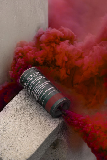 Red smoke grenade resting on a concrete ledge, dense crimson smoke billowing upward against a wall corner