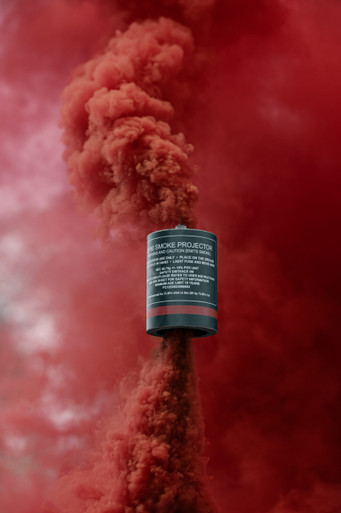 Red smoke grenade with a tall, dense red smoke column rising straight up against a blurred red backdrop