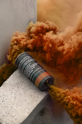 Orange smoke grenade resting on a concrete ledge, amber-orange smoke billowing upward against a wall corner