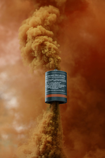 Orange smoke grenade with a tall, dense amber-orange smoke column rising straight up against a blurred backdrop