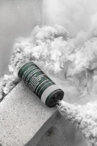 White smoke grenade resting on a concrete ledge, dense white smoke billowing upward against a wall corner