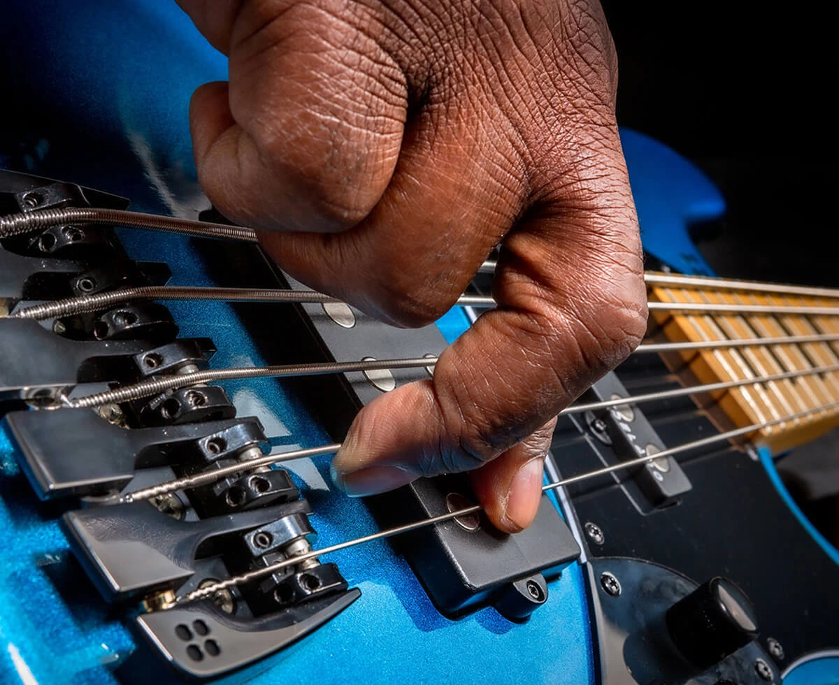 A player's hand plucking the strings of a five-string bass near the bridge.