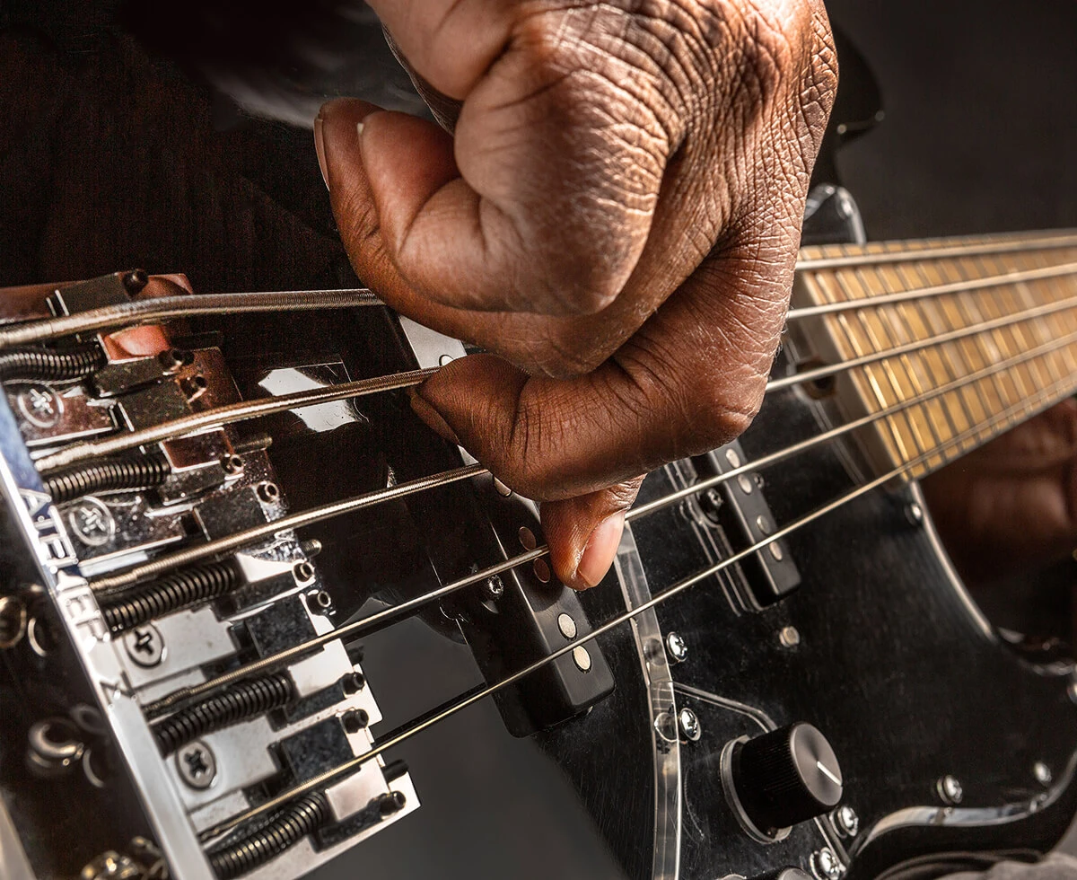 A player's hand plucking the strings of a five-string bass near the bridge.