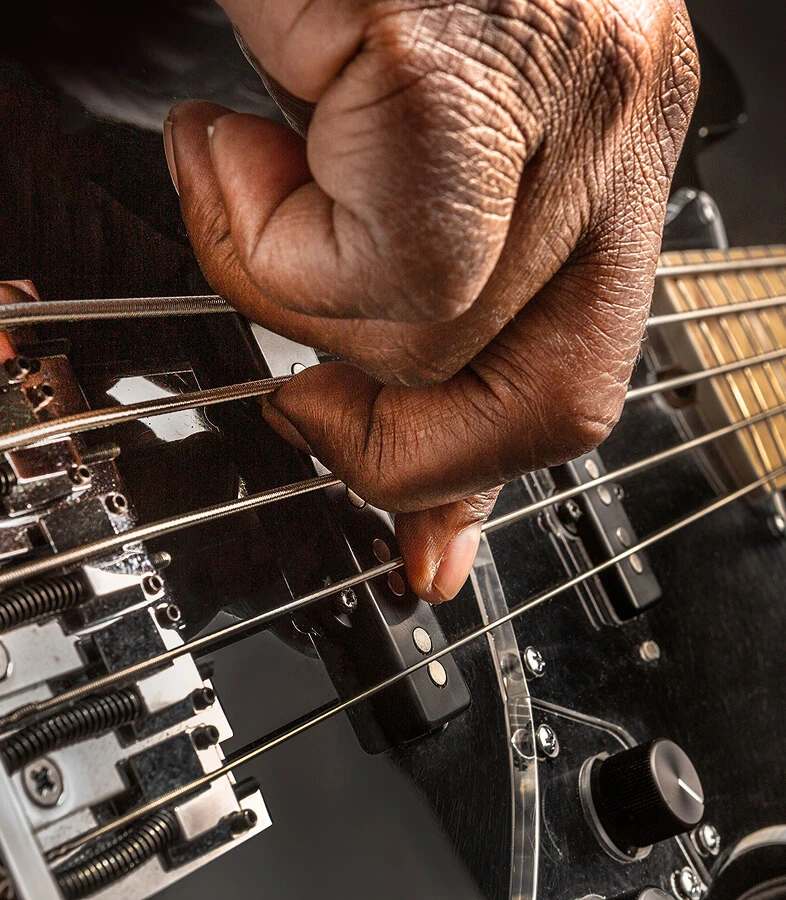 A player's hand plucking the strings of a five-string bass near the bridge.