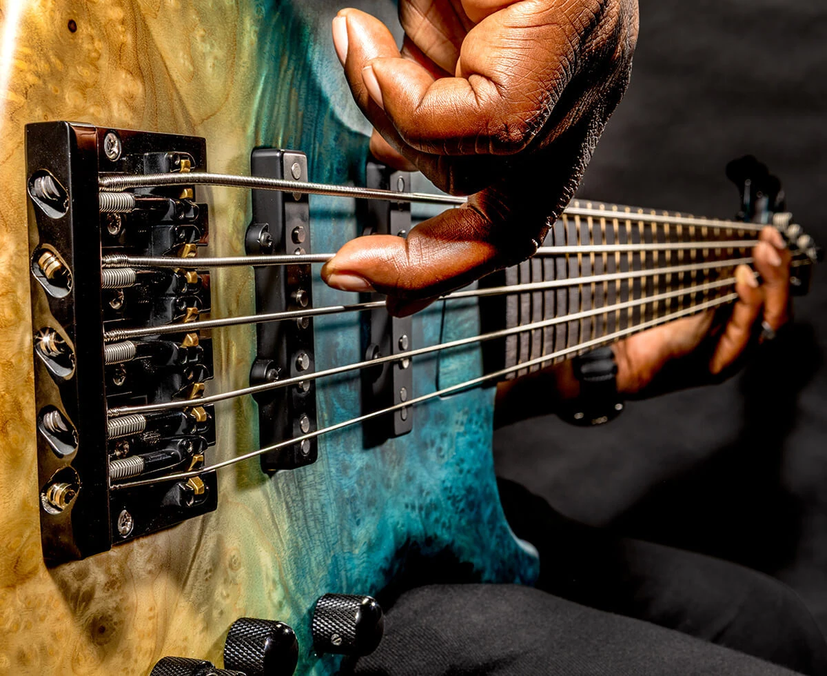 A player's hand plucking the strings of a five-string bass near the bridge.
