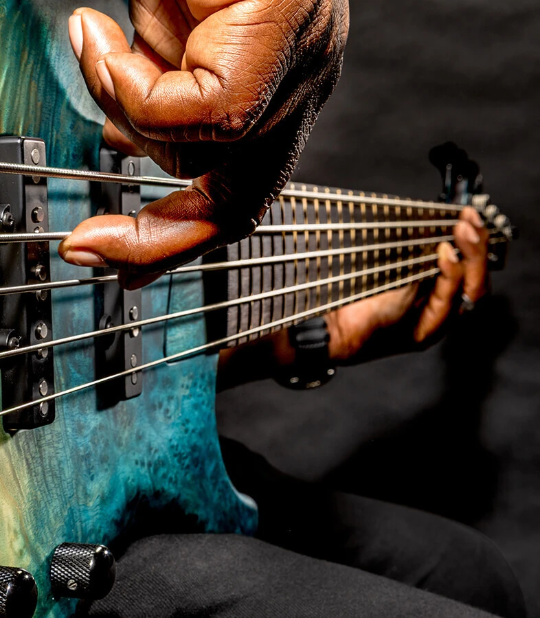 A player's hand plucking the strings of a five-string bass near the bridge.
