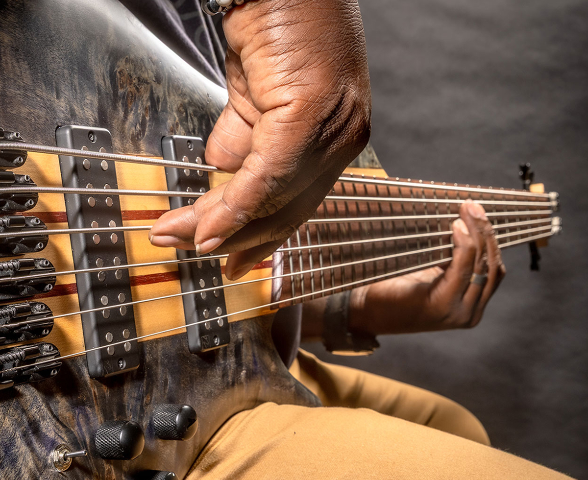 A player's hand plucking the strings of a six-string bass near the bridge.