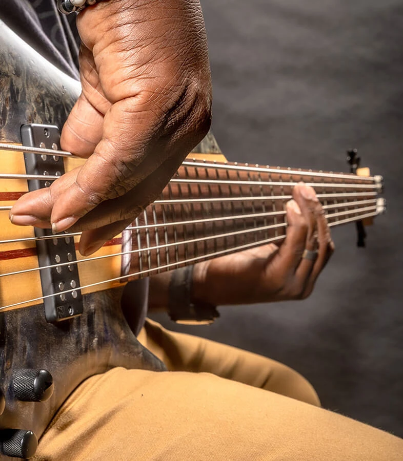 A player's hand plucking the strings of a six-string bass near the bridge.