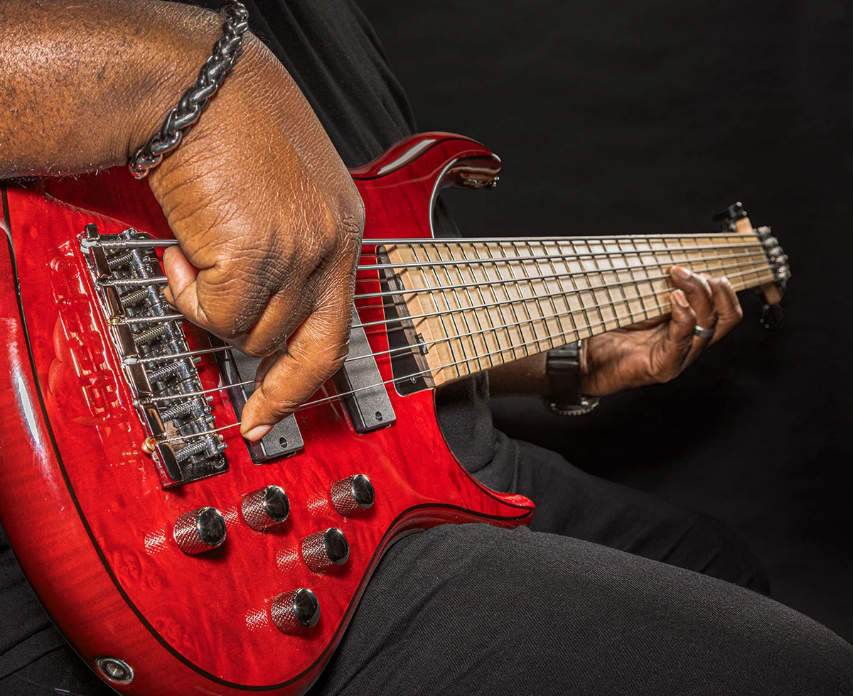 A player's hand plucking the strings of a six-string bass near the bridge.