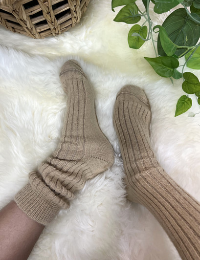 A person's feet wearing ribbed midcalf knitted socks in light brown color on a fluffy white sheepskin rug.
