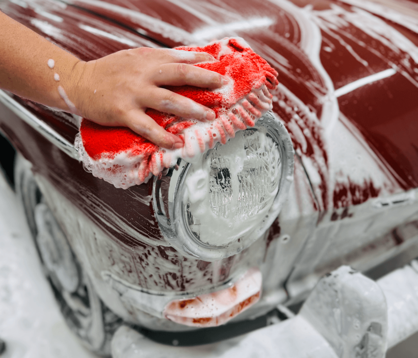 Man washing the headlight of a classic car with a red wash sponge in heavy foam.