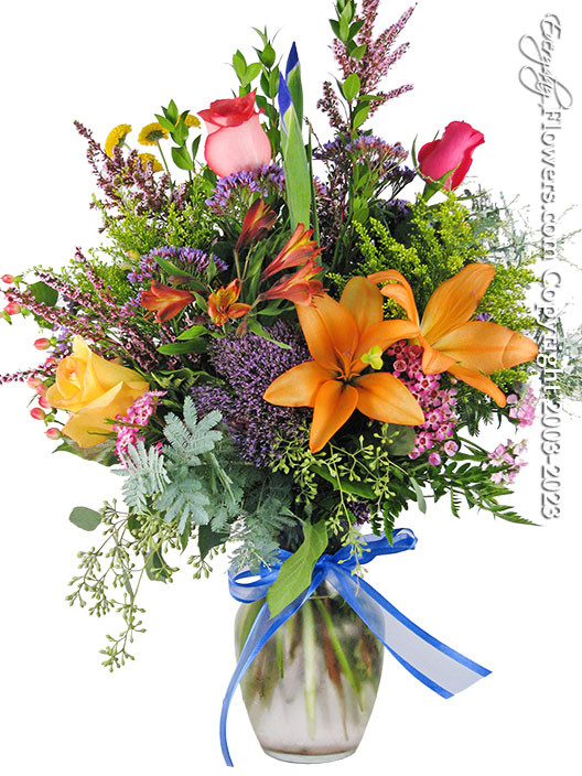 Orange Tiger Lilies, Pink, Red and Yellow Roses, Alstromeria, Yellow Chrysanthemum Buttons, Blue Iris, Assorted Seasonal Fillers, and A Blue Hand Tied Silk Ribbon arranged in a  8 Inch Ginger Glass Vase.