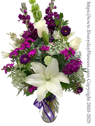 Purple and white flowers arranged in a glass vase using white lilies, white roses, purple hoary stock, purple asters or purple lisianthus, purple alstroemeria and a purple bow tied around the vase