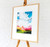 Bright landscape art print framed in a warm oak frame, displayed on a wooden easel.