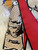 The image shows a Louis Vuitton scarf with a distinctive design. The scarf features a combination of red and beige colors, with the beige sections adorned with the classic Louis Vuitton monogram pattern. The design also includes artistic illustrations of what appear to be Louis Vuitton bags. The brand name "LOUIS VUITTON" is prominently displayed on the red section of the scarf. The scarf is placed on a white fur surface, with a Louis Vuitton box and a receipt partially visible in the background