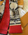 The image shows a Louis Vuitton scarf with a distinctive design. The scarf features a combination of red and beige colors, with the beige sections adorned with the classic Louis Vuitton monogram pattern. The design also includes artistic illustrations of what appear to be Louis Vuitton bags. The brand name "LOUIS VUITTON" is prominently displayed on the red section of the scarf. The scarf is placed on a white fur surface, with a Louis Vuitton box and a receipt partially visible in the background