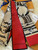 The image shows a Louis Vuitton scarf with a distinctive design. The scarf features a combination of red and beige colors, with the beige sections adorned with the classic Louis Vuitton monogram pattern. The design also includes artistic illustrations of what appear to be Louis Vuitton bags. The brand name "LOUIS VUITTON" is prominently displayed on the red section of the scarf. The scarf is placed on a white fur surface, with a Louis Vuitton box and a receipt partially visible in the background