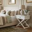 Hampstead Cross Leg Bedside Side Table in white styled beside a contemporary bed with a ceramic lamp and decorative greenery in a light-filled coastal-inspired bedroom.